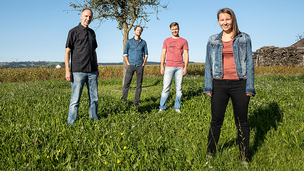 Lara Amalia steht mit ihren Band-Kollegen Gunter, Andreas und Clemens in der Sonne in der Wiese. Dahinter sieht man einen Baum und einen Holzhaufen.