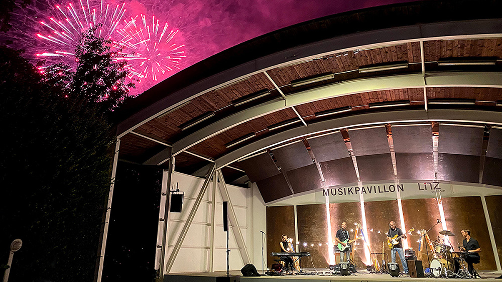 Lara Amalia mit Band auf der Bühne im Musikpavillon Linz. Dahinter ein Feuerwerk.